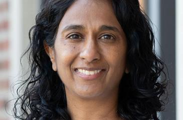 A portrait of Shalini smiling facing the camera. She wears a light blue shirt with a scoop neck hem. Her curly black hair is resting on the tops of her shoulders.