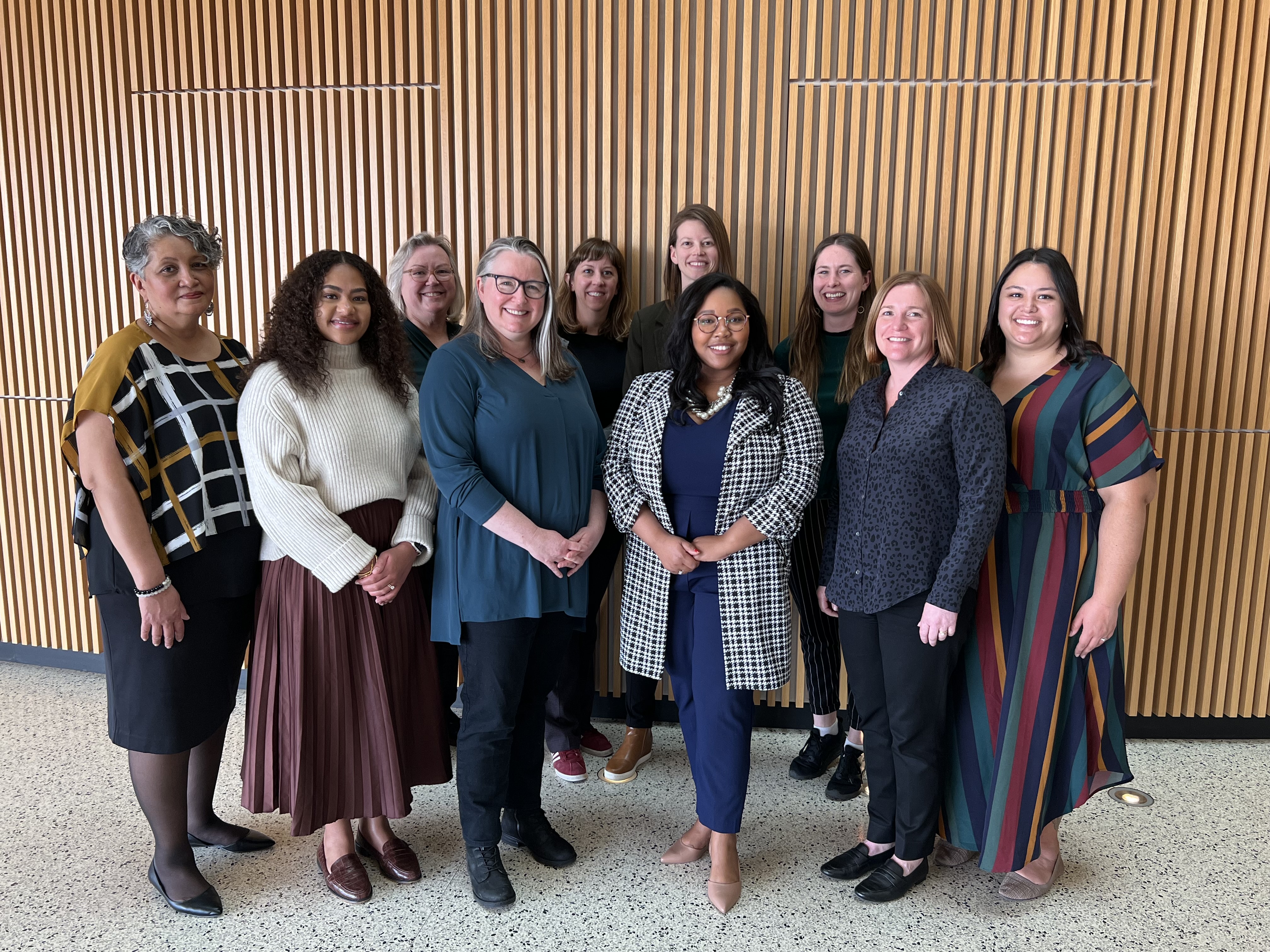 The MCC community outreach and engagement team poses in front of an oak-colored wood panel wall on the UMN Twin Cities campus. There are 10 women in the photo, standing with hands clasped or at their sides.