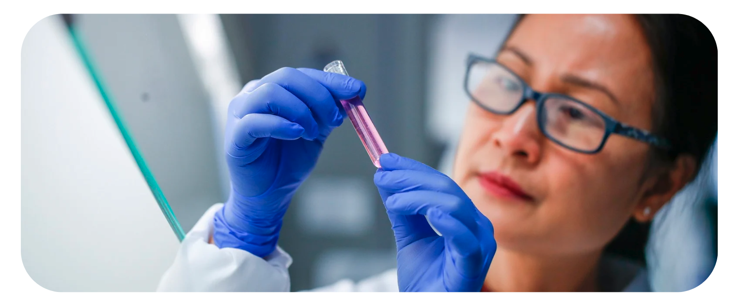 A woman of Asian descent wearing black framed glasses with her black hair pulled back into a neat ponytail is wearing blue exam gloves, a white coat, and studying a test tube in front of her filled with a pinkish-red liquid.