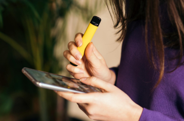 A person, presumed to be a teenager, with long hair wearing a purple long sleeve shirt holds in their right hand a yellow vaping pen and in their left hand a smartphone. 