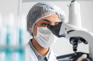 A close up of a female researcher working on a microscope. The researcher wears a white hair net and white mask over the bottom half of her face. 