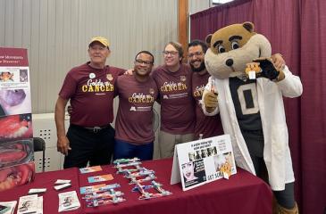 A group of men wearing maroon shirts with the "Goldy vs. Cancer" logo on them pose for a photo next to Goldy Gopher. They are all smiling and standing together behind a display table with a maroon tablecloth at the Minnesota State Fair.